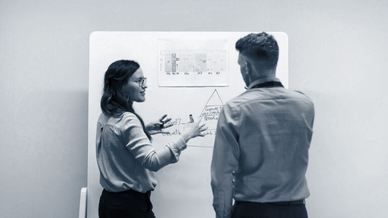 Two business professionals collaborate in front of a whiteboard, reviewing marketing workflows and strategic planning to improve campaign performance and jump-start stalled growth.